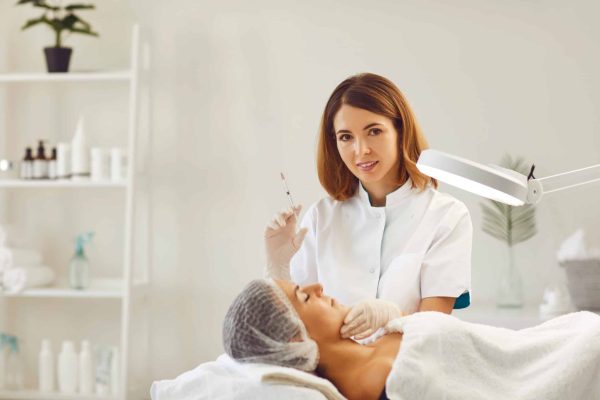 Young smiling woman cosmetologist looking at camera and ready to make facial beauty injection for lying woman in beauty spa salon. Facial treatment, skincare, cosmetology concept